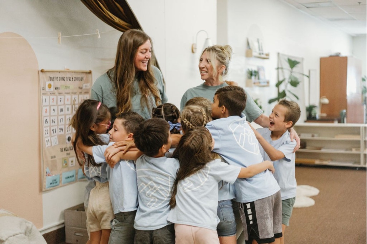 Two laughing teachers in a big group hug with kids in light-blue Legacy shirts, classroom calendar board behind them