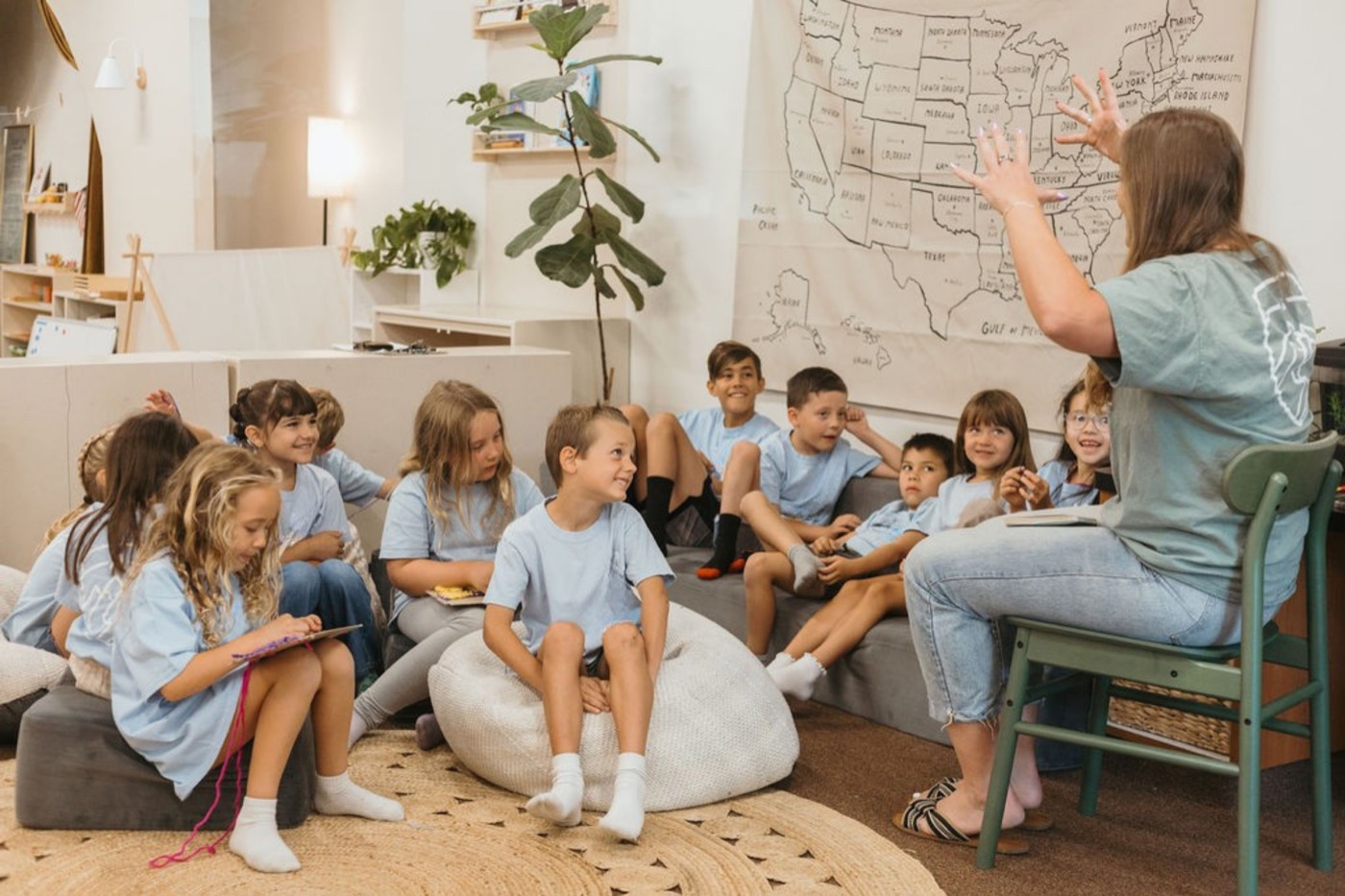 Teacher (seen from behind) tells a story with big hand gestures to kids in light-blue shirts on bean bags and a couch, large US map on the wall